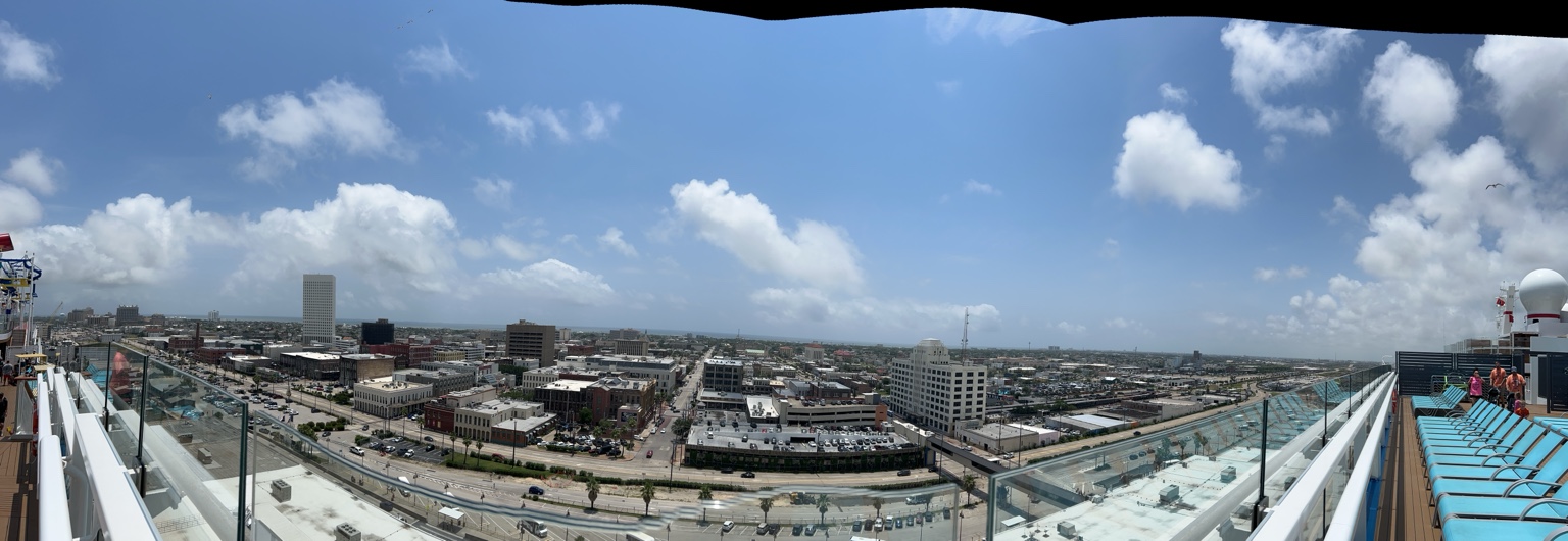 Wide city and harbor panorama seen from a ship deck beneath a bright, cloud-dotted sky.