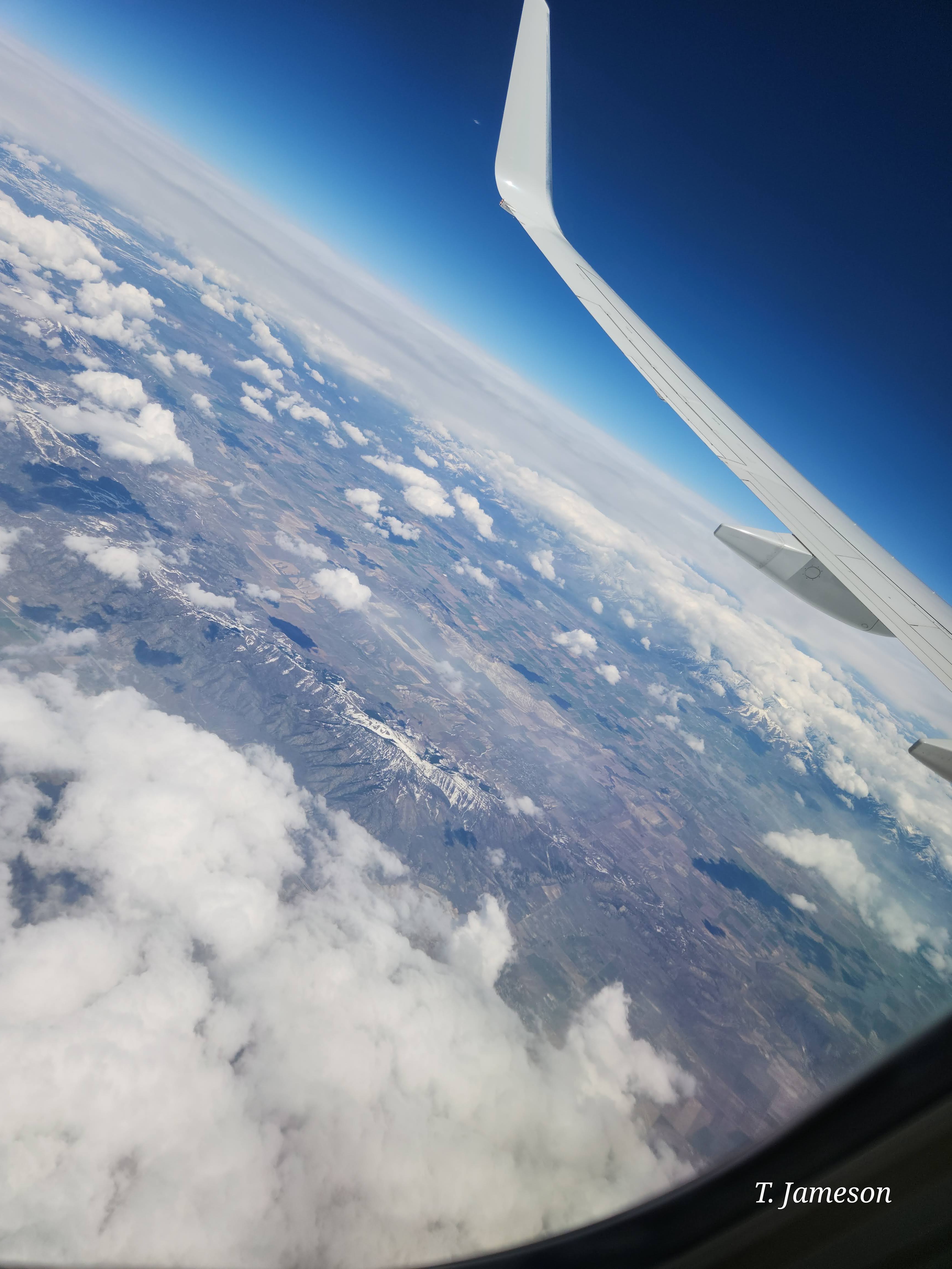 View from an airplane window across clouds and a mountain landscape beneath a blue sky.