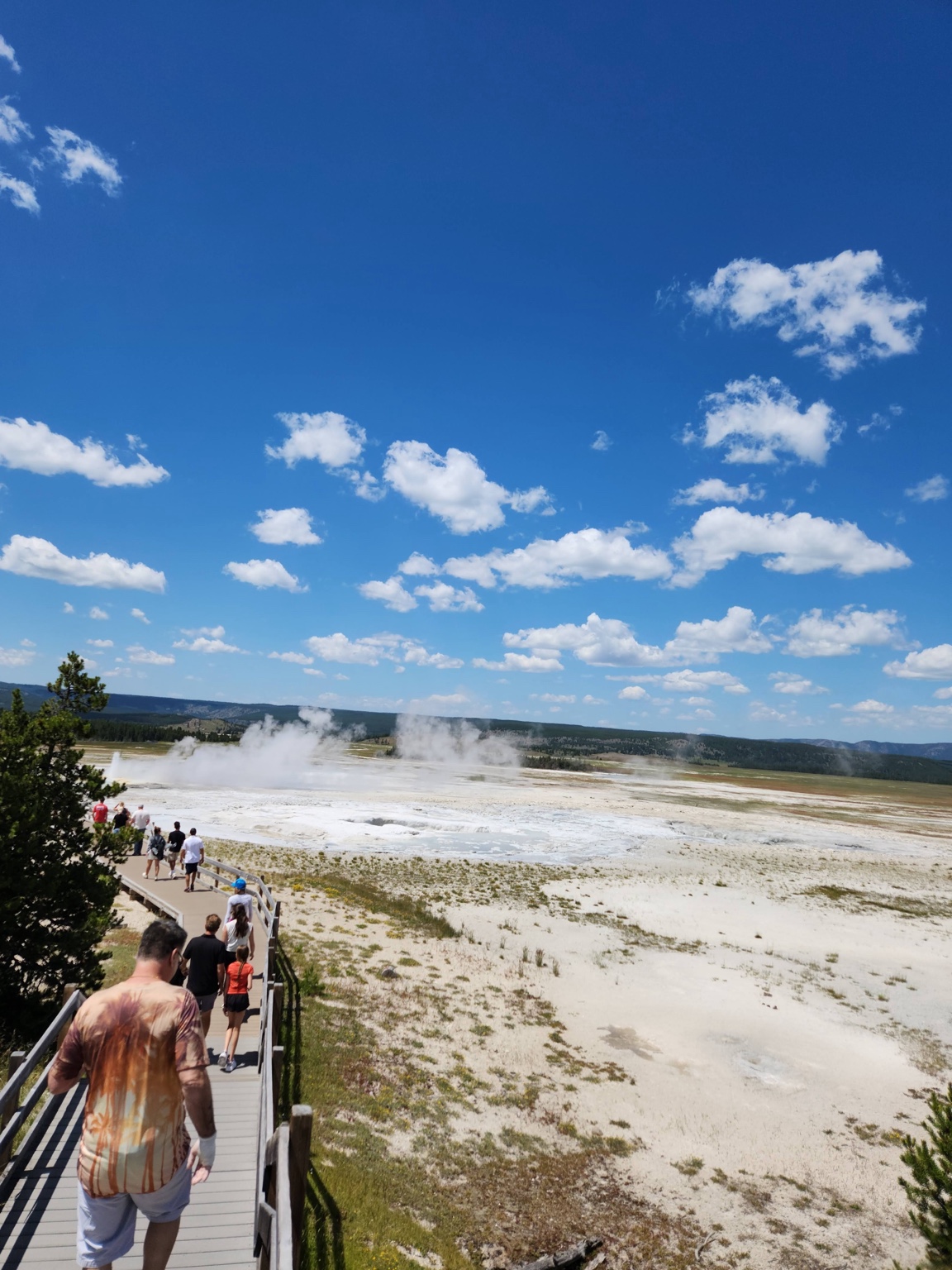 Visitors walking along a boardwalk through a pale geothermal landscape under scattered clouds.
