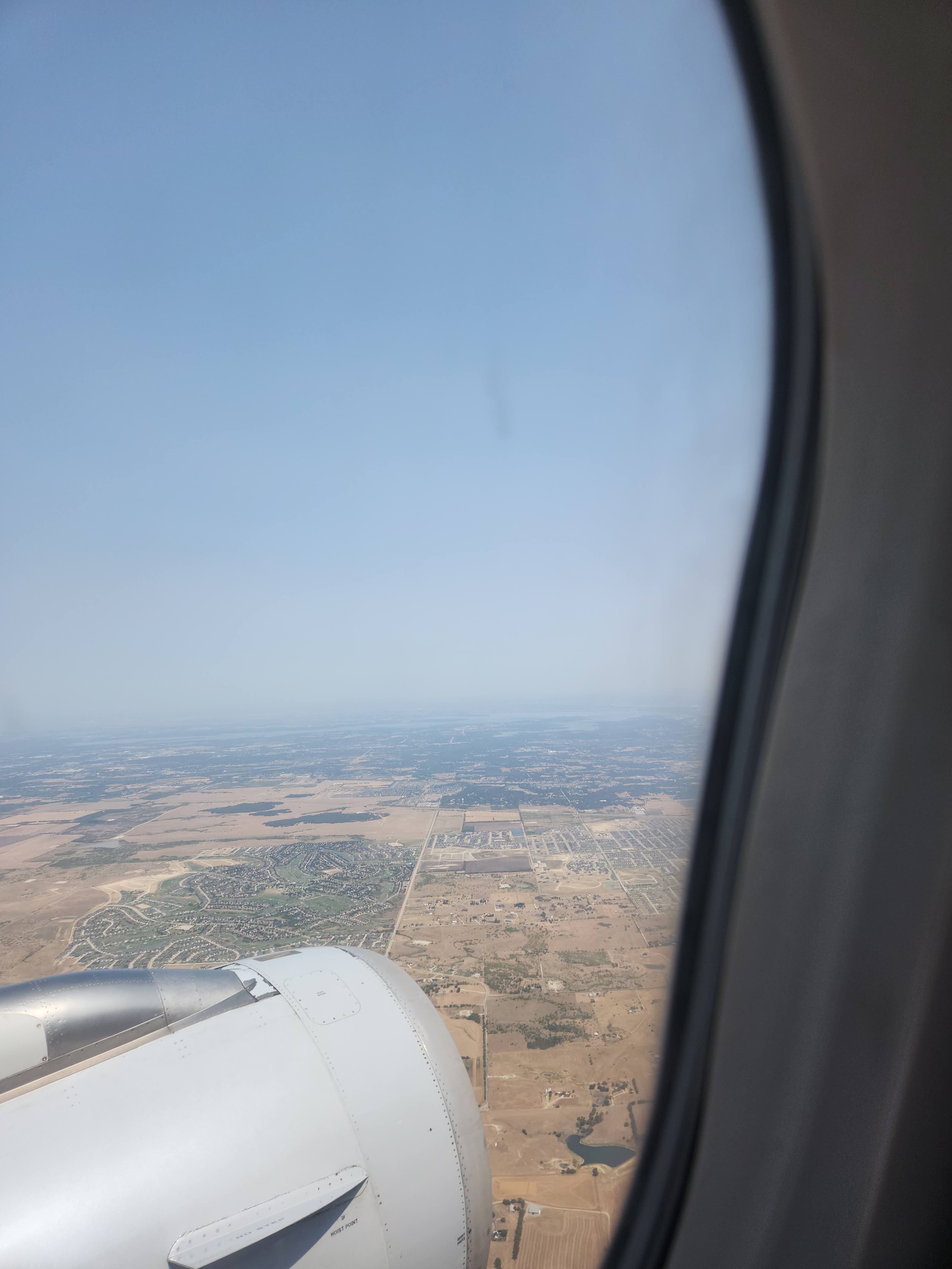 Airplane wing and engine above a hazy patchwork of fields, roads, and neighborhoods.