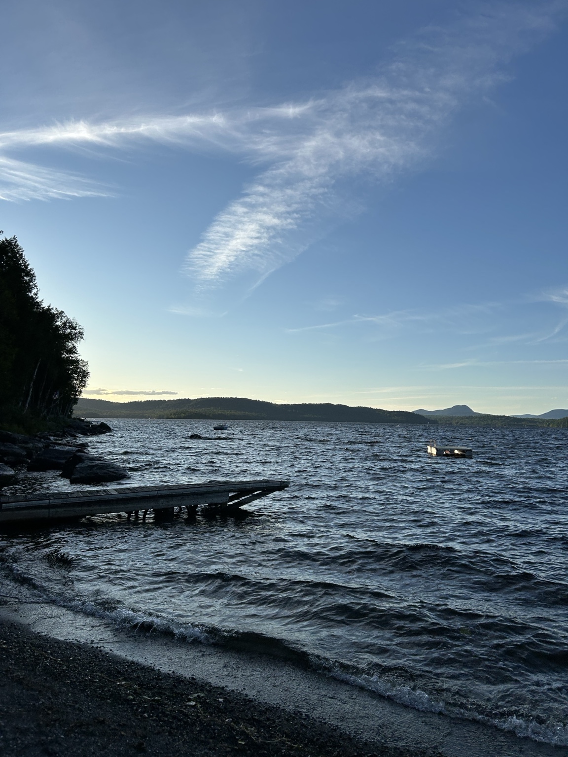 Wind-textured lake water beneath a blue sky, with a dock, wooded shoreline, and distant hills.