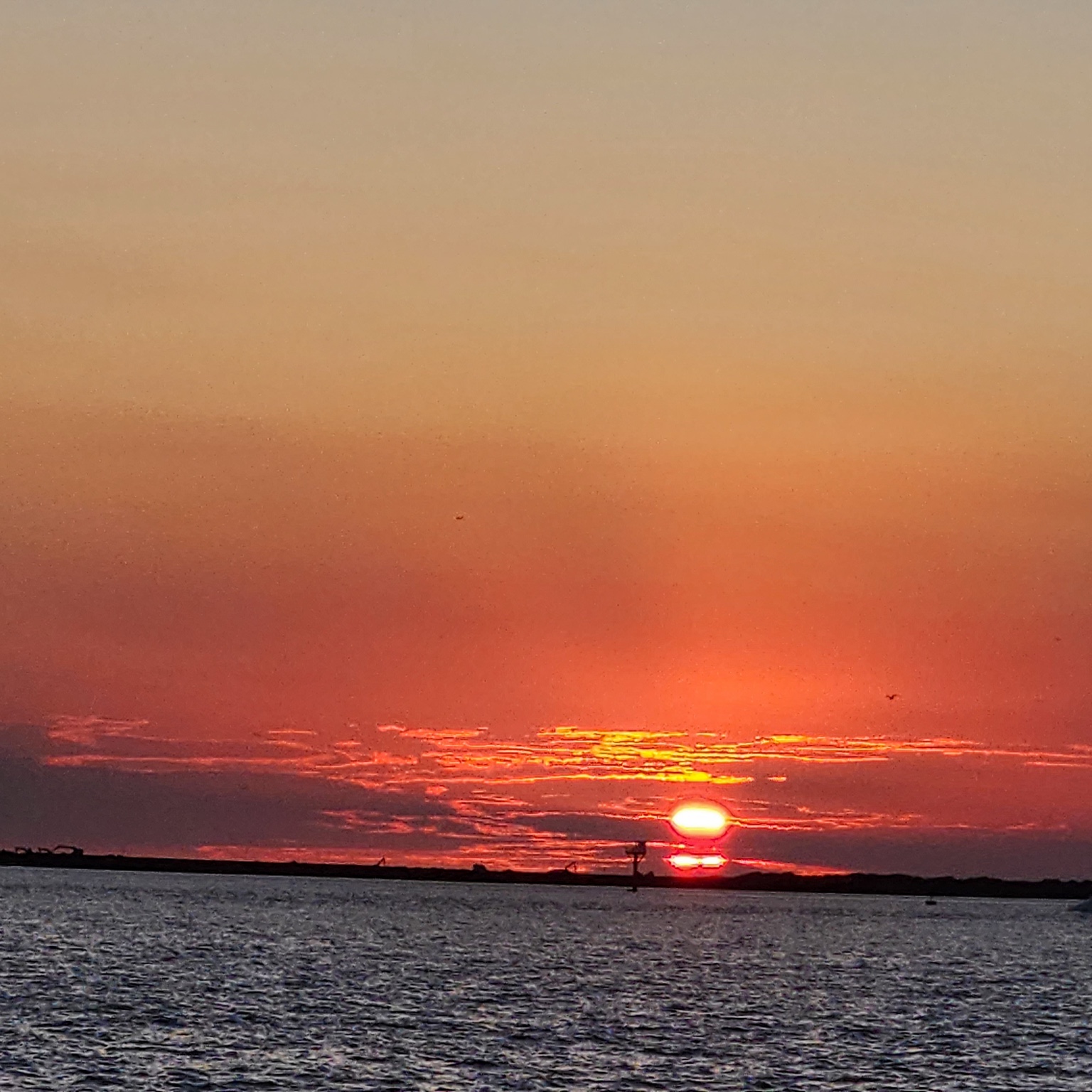 Bright orange sun setting at the horizon over rippled open water, with vivid red-orange clouds.